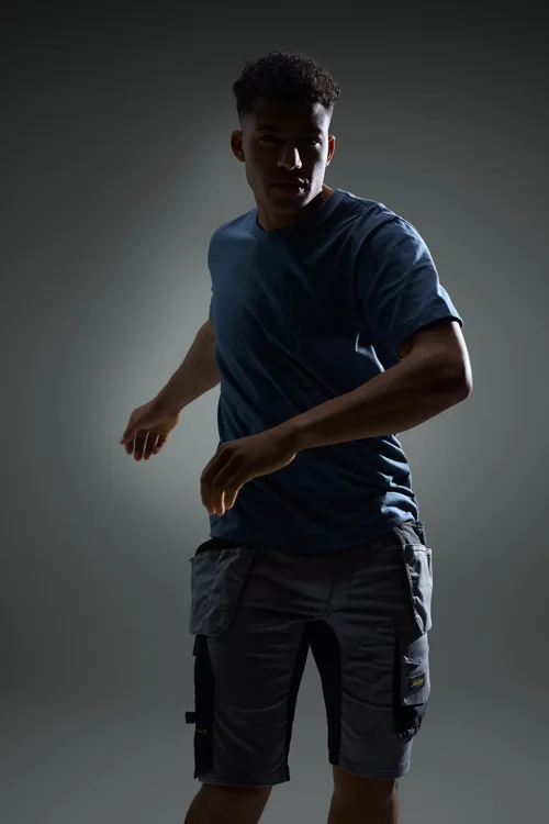 Young man in a blue t-shirt and gray cargo shorts strikes a dynamic pose against a dark background in a studio.