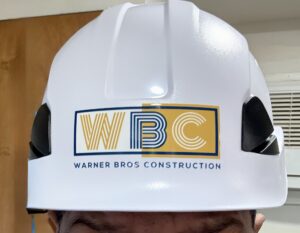 Close-up of a white hard hat with a yellow-and-blue Warner Bros Construction logo on the front, obscuring most of the wearer's face.