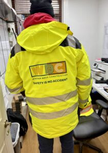 Back view of a construction worker in a neon yellow safety jacket with reflective stripes and a red hoodie, inside a cluttered office space.