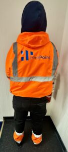 Person in a bright orange high-visibility jacket with reflective strips and 'HirePoint' logo, standing with back to camera in a hallway.