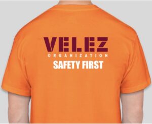 Back view of a person wearing a bright orange T-shirt with a bold maroon 'VILEZ Organization' logo and a white 'SAFETY FIRST' slogan.