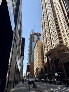 Low-angle view of a city street with tall glass and brick buildings on either side and a construction crane against a clear blue sky in the distance